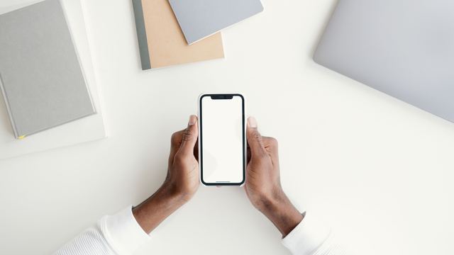 Two hands holding a smartphone with a blank screen above a white desk with notebooks and a closed laptop.