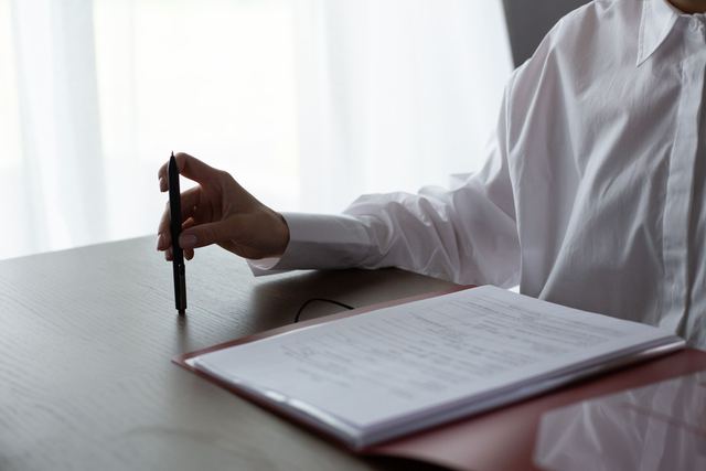 A person holding a pen beside an open folder of printed papers on a wooden table, face out of frame.