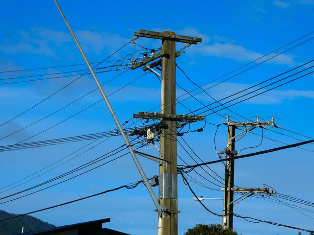 Concrete power poles with crossarms, insulators and a web of overhead lines against a blue sky, with bush-covered hills behind.