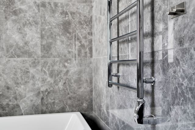 A chrome electric heated towel rail on a grey marble-tiled bathroom wall beside a bath.
