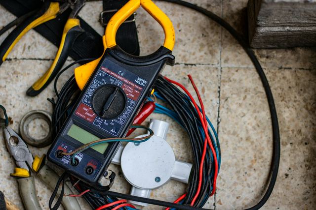 A clamp meter lying on coiled red, blue and black wires beside pliers and a white junction box on a tiled floor.