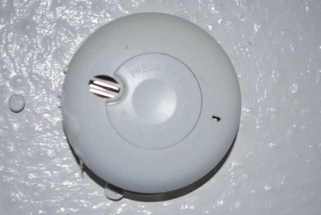 Close-up of a ceiling-mounted smoke alarm with a press-to-test button, fixed to a textured ceiling.